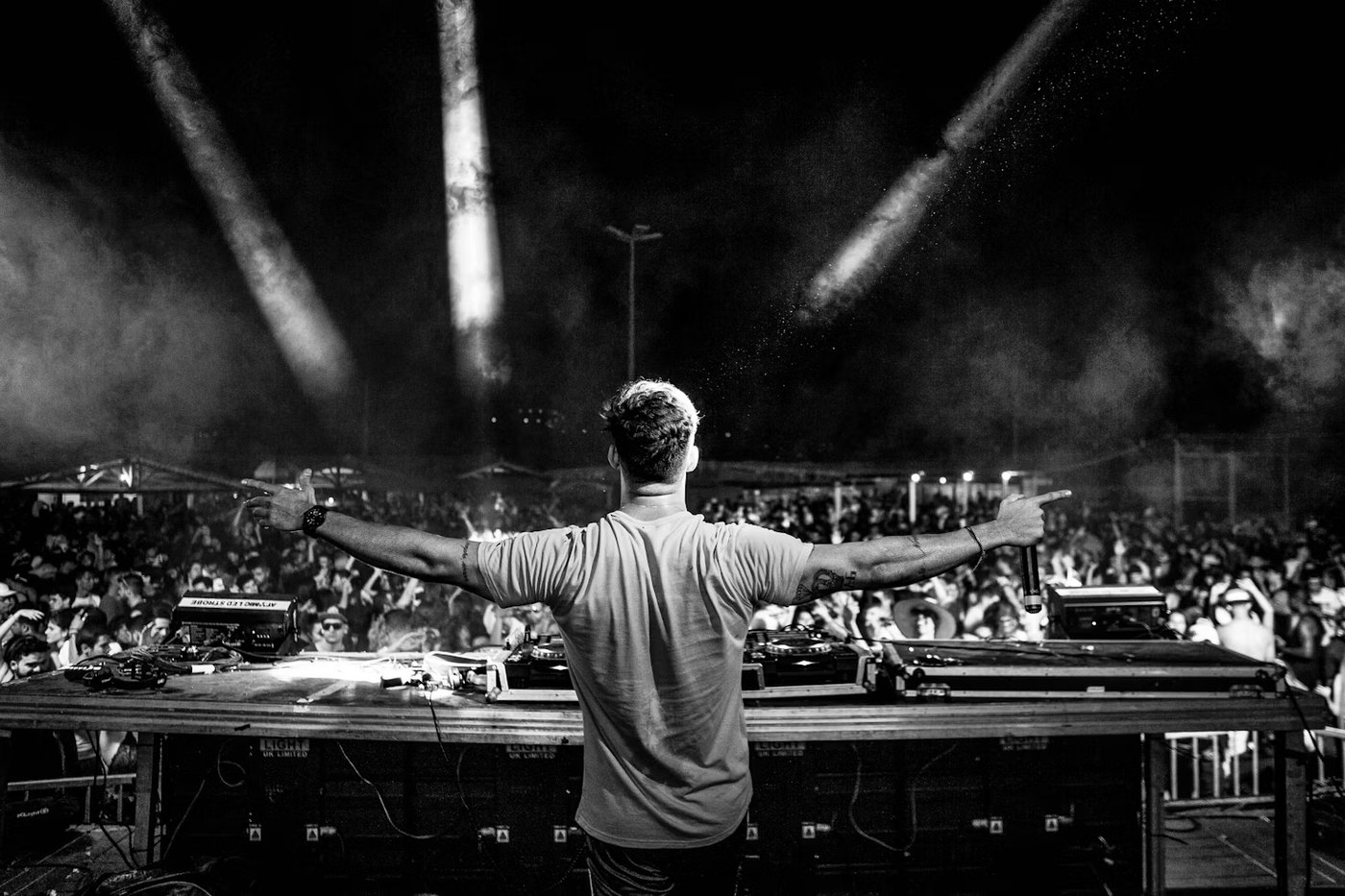 A DJ facing a festival crowd from behind the decks, arms out, spotlights overhead